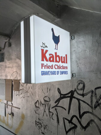 Im Treppenhaus � wie an anderen Orten in Kassel � l�sst sich eine Arbeit von Hamja Ahsan entdecken. Seine Leuchtschilder machen Werbung f�r eine Franchise-Kette konkurrierender H�hnchenbrater. Die fiktiven Tafeln setzen sich mit islamophoben Klichees und Stereotypen auseinander.